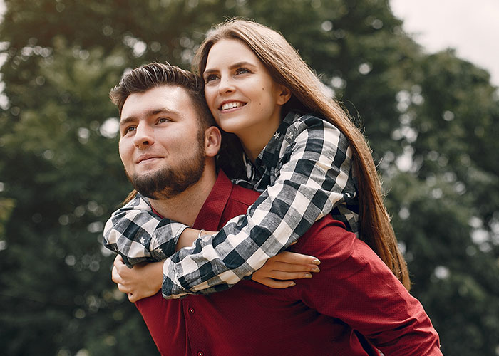 A couple in a playful piggyback pose, with the man in a red shirt and the woman in a checkered shirt, outdoors.