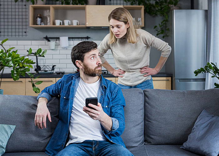 Man on sofa with phone, looking surprised as woman confronts him, illustrating temptation and conflict.