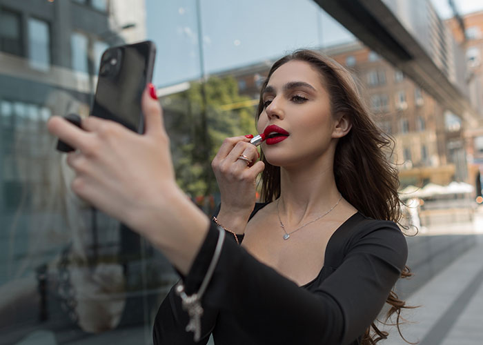 Woman applying lipstick while taking a selfie, symbolizing temptation in a city setting.
