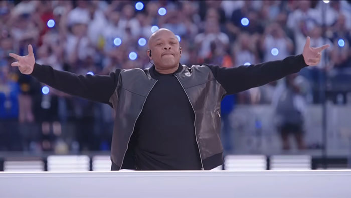 Talented person on stage spreading arms wide, wearing a leather jacket, with a crowd and lights in the background.