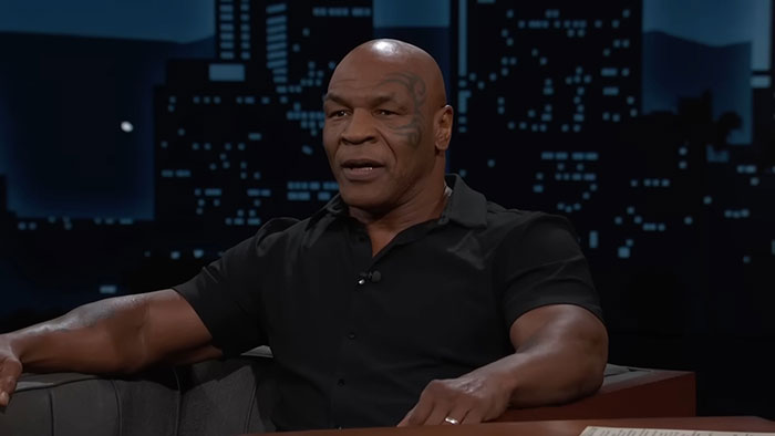 A person sitting on a talk show set, wearing a black shirt, engaged in conversation about talented people.
