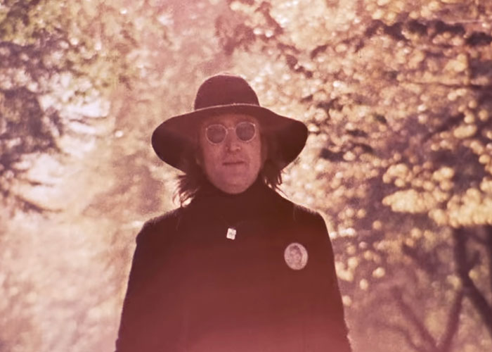A person in a hat and glasses walks through a sunlit forest scene.