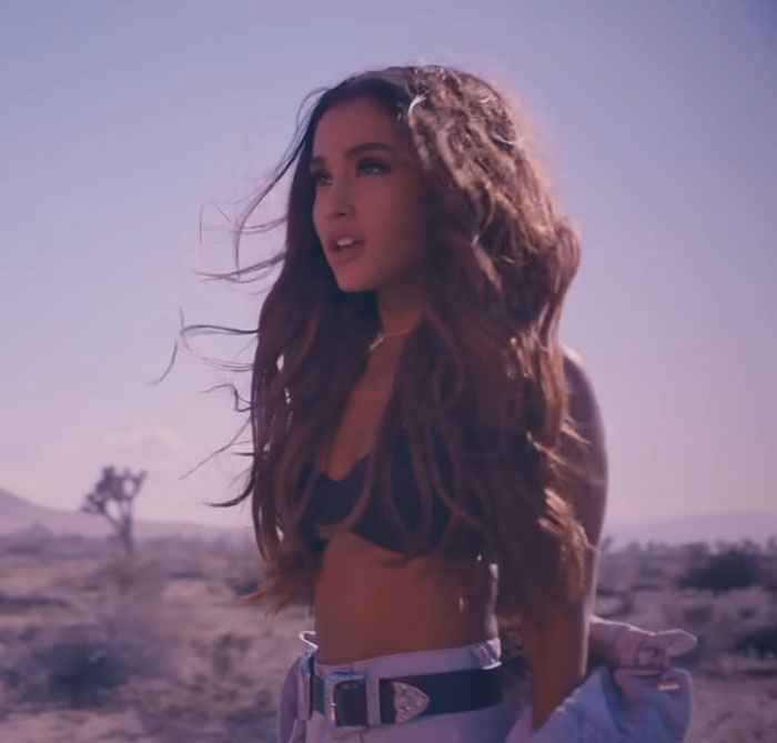 Young woman in a desert, wearing a black top and unbuttoned denim. Long hair blowing in the wind under a clear sky.