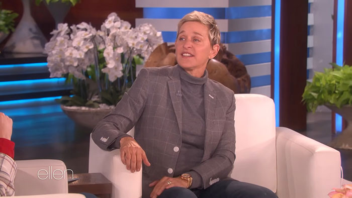 Celebrity on a talk show, wearing a gray suit jacket and turtleneck, seated in a white chair.