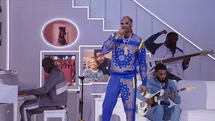 Musician in blue outfit performing with a band, featuring keyboard, bass, and electric guitar under framed photos.