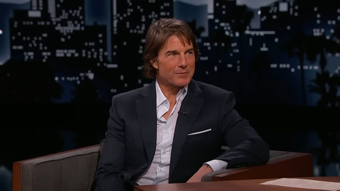 “Celeb in a suit on a talk show set, discussing topics under city night skyline backdrop.”