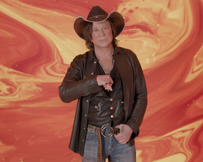 Person in leather jacket and cowboy hat stands against a vibrant orange background.
