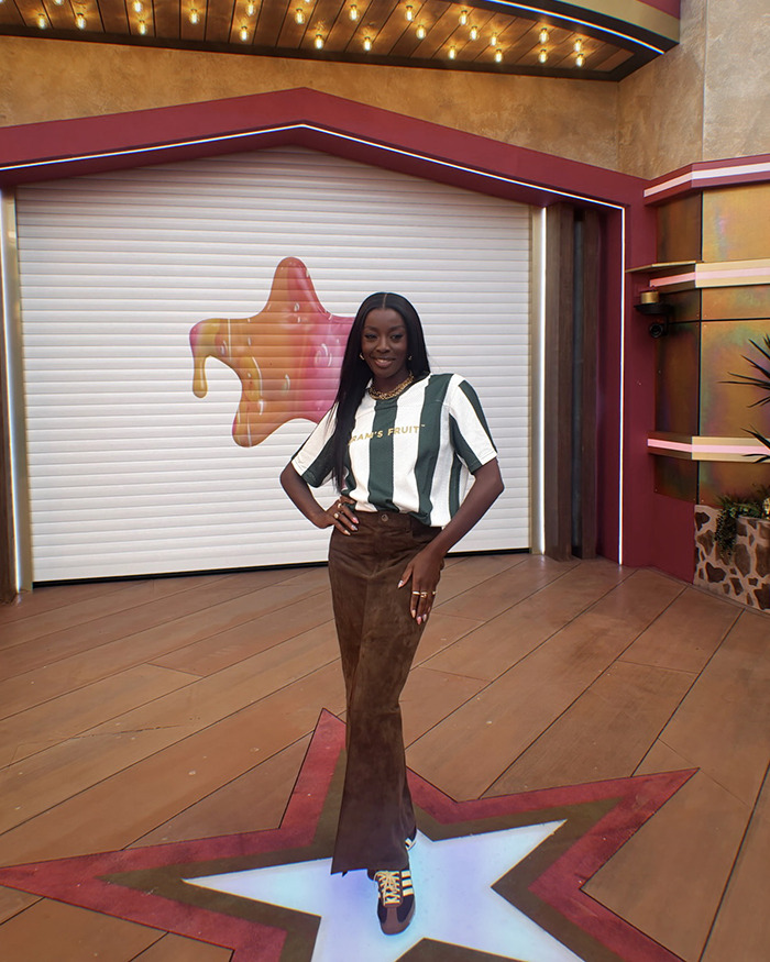 AJ Odudu smiling in a striped shirt and brown pants on the set of Celebrity Big Brother.