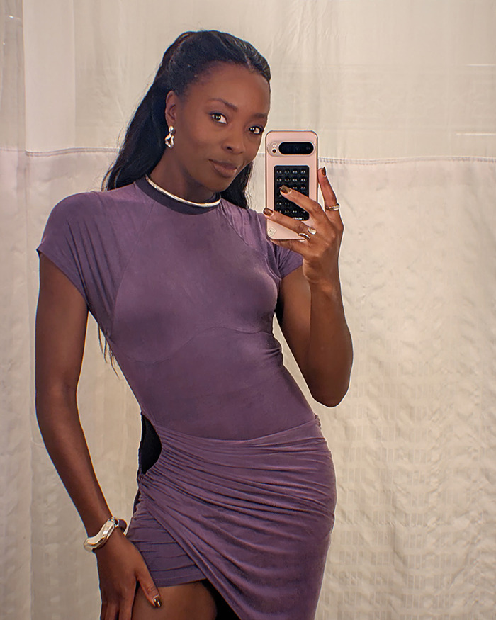 AJ Odudu wearing a purple dress, holding a smartphone, posing for a mirror selfie.