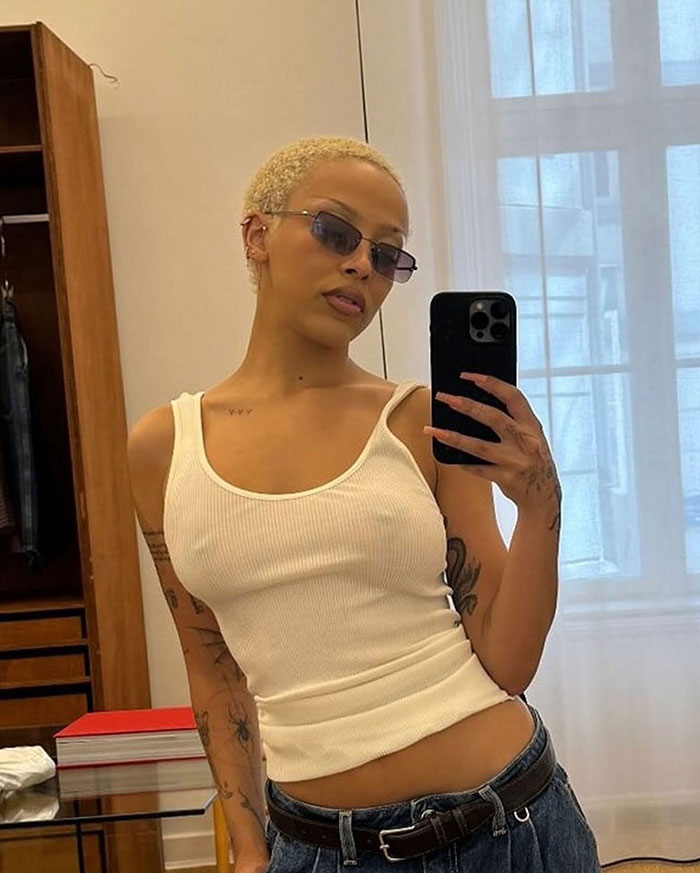 Young person in sunglasses and a white tank top taking a mirror selfie, showcasing style and confidence.