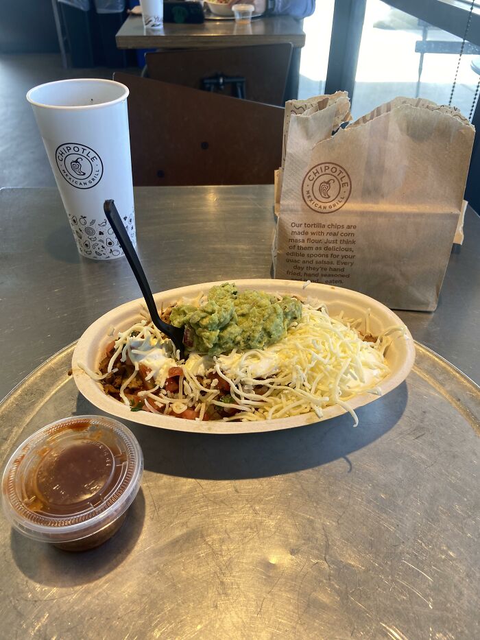 Chipotle bowl with guacamole and cheese in a chain restaurant setting, next to a cup and paper bag.