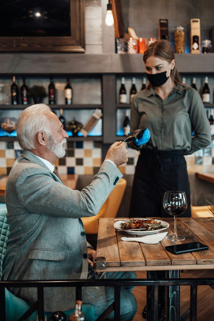 Elderly man in suit pays with card, served by waitress in mask at chain restaurant.