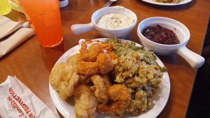 Plate of fried seafood, stuffing, and green beans from a chain restaurant, with sauces and orange drink nearby.