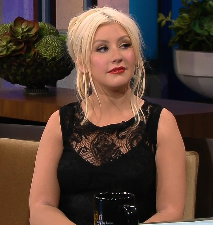Christina Aguilera during an interview wearing a black lace top with blonde hair styled up and red lipstick.