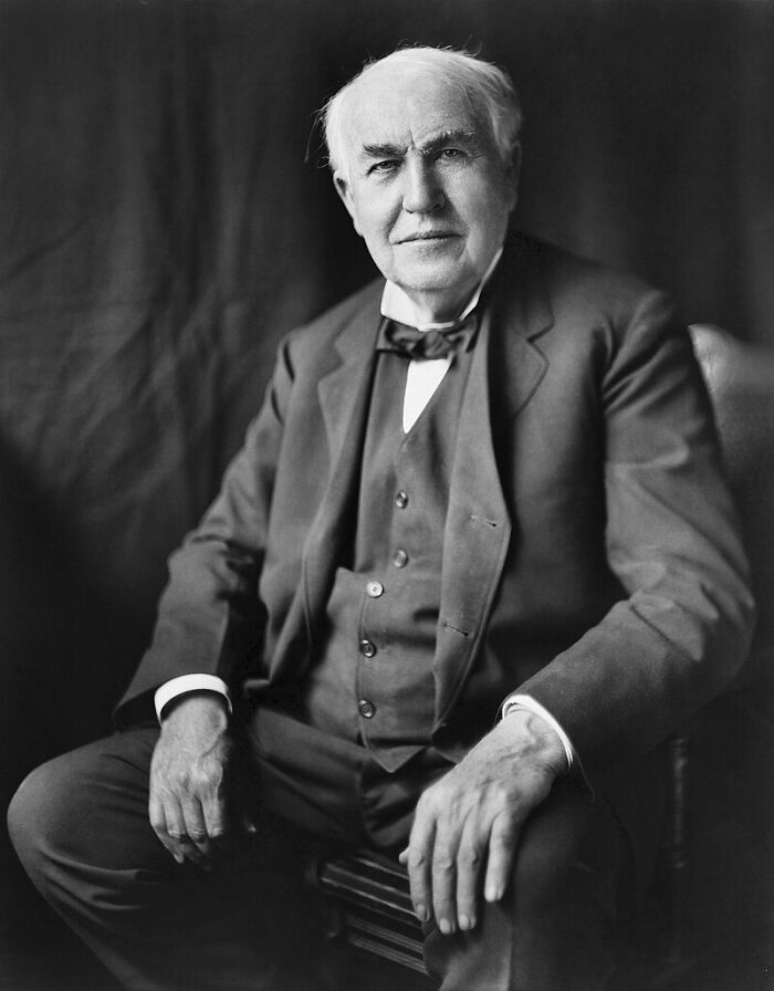 A distinguished elderly man in a formal suit, seated and looking into the camera, embodying infamous historical figures.