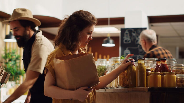 A woman in a deli, holding a paper bag, picks a jar from the shelf.
