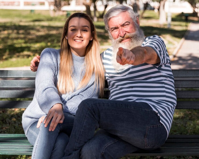 Smiling man and woman sitting on a park bench, man pointing toward the camera, outdoors.