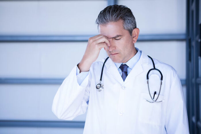 Doctor in white coat, looking stressed, with stethoscope, indoors, conveying questions burden.