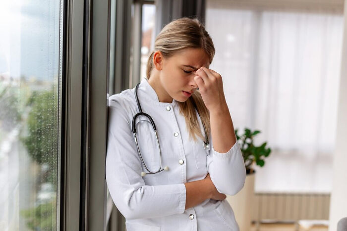 Doctor in white coat looking stressed, standing by a window with a stethoscope around her neck.