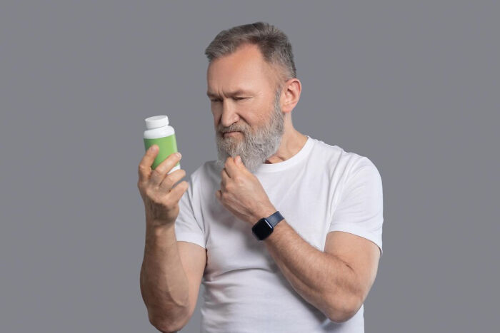 Man examining a pill bottle, pondering questions doctors often hear from friends, wearing a white shirt.