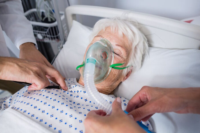 Elderly patient with oxygen mask in hospital bed, attended by doctors.
