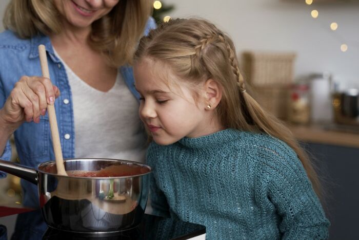 Girl in a blue sweater, playfully sniffing a cooking pot, embodying child comedians' innocent curiosity.