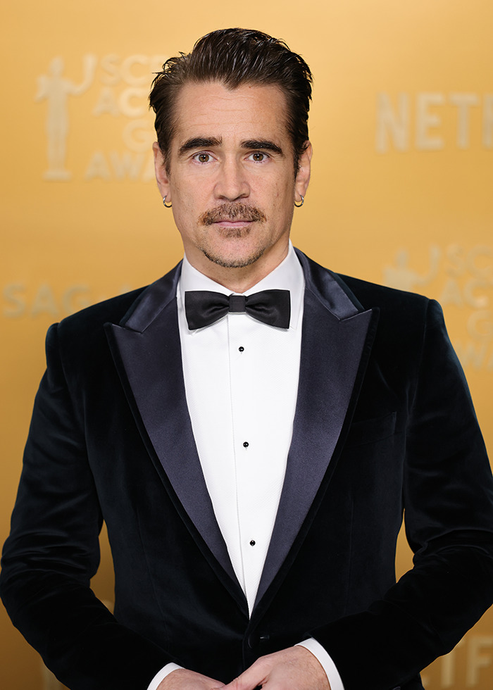 Actor in a black tuxedo with a bow tie at an awards event, standing against a gold backdrop.