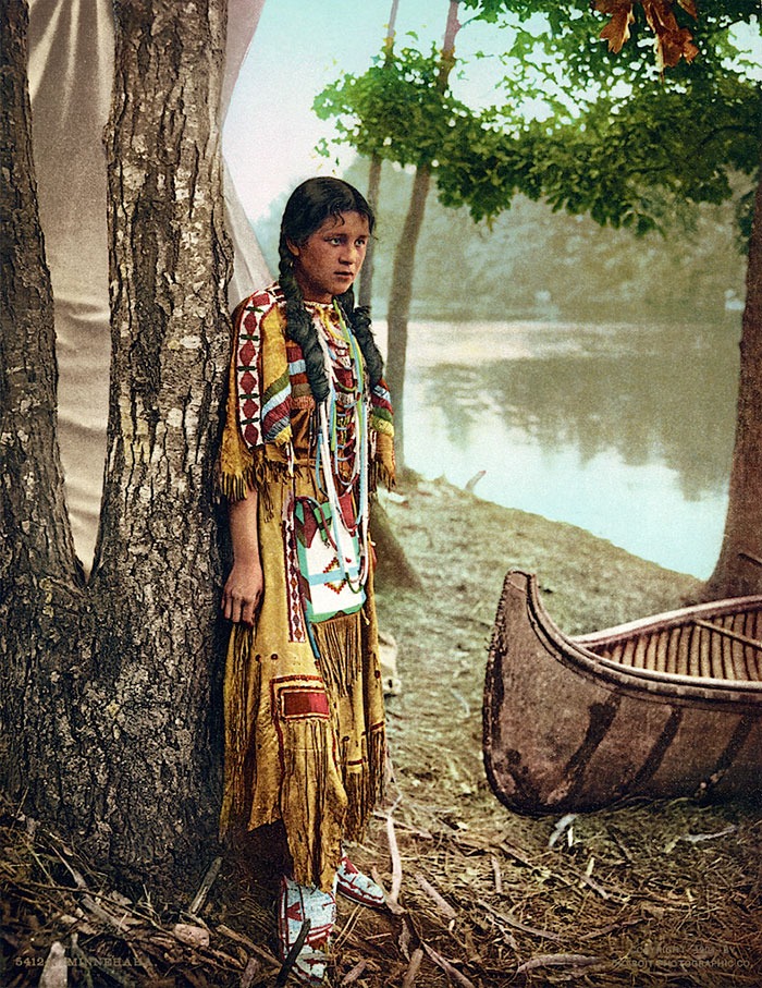 Young Native American woman in traditional dress standing by a tree and canoe by the water in a historic photo.