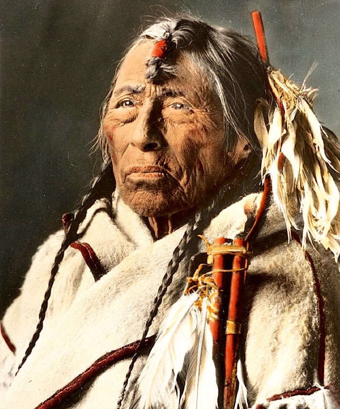 Elder Native American man wearing traditional clothing and holding a decorated staff in a haunting historic portrait.