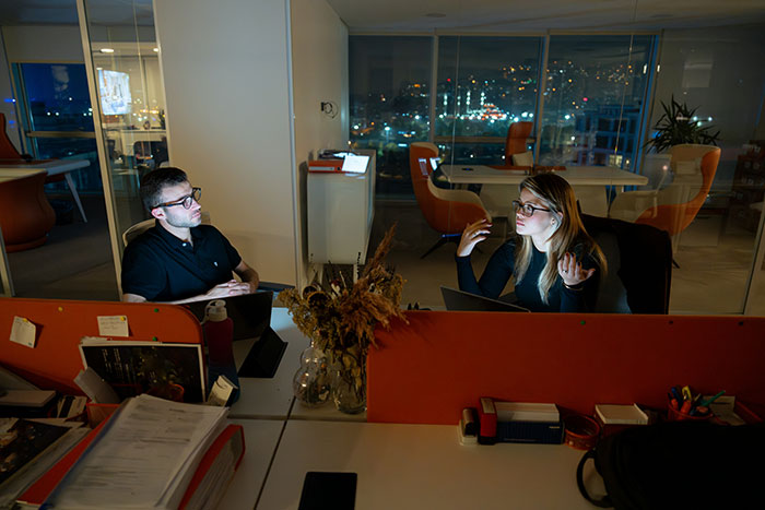 Two employees discussing work perks in a modern office at night, illuminated by laptop screens.