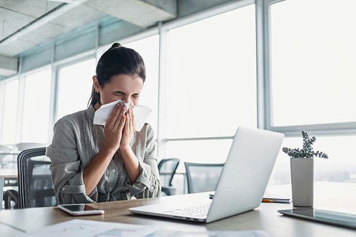 Employee using a tissue at a desk in a bright office, highlighting work perks for well-being.