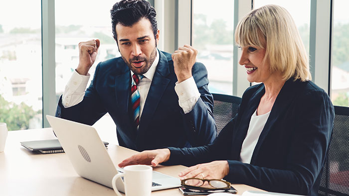 Two employees in a meeting room celebrate a successful presentation; a laptop and coffee cups are on the table.