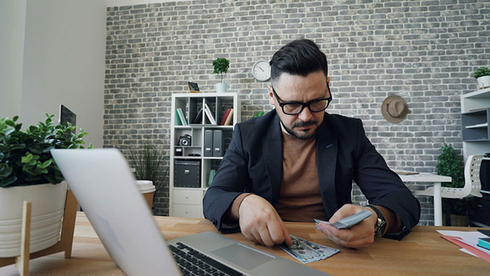 Man in office, counting money at desk, reflecting employee work perks in a modern workspace with a laptop and bookshelves.