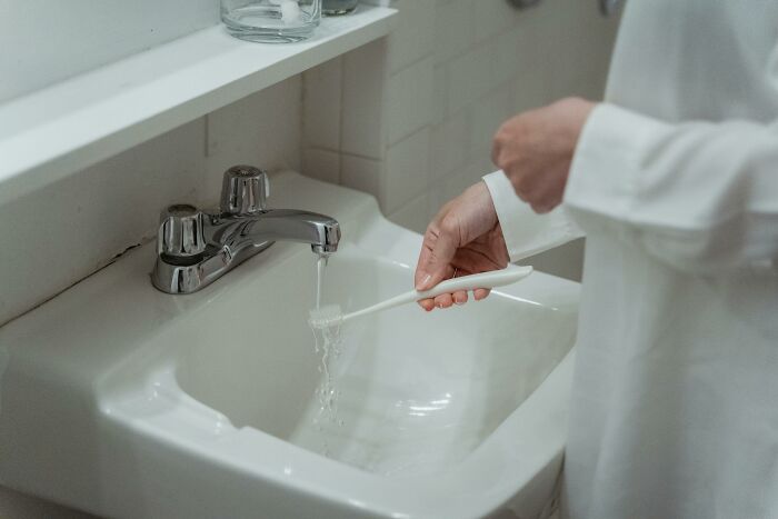 Person in white rinsing a toothbrush under a faucet, possibly pondering conspiracy theories.