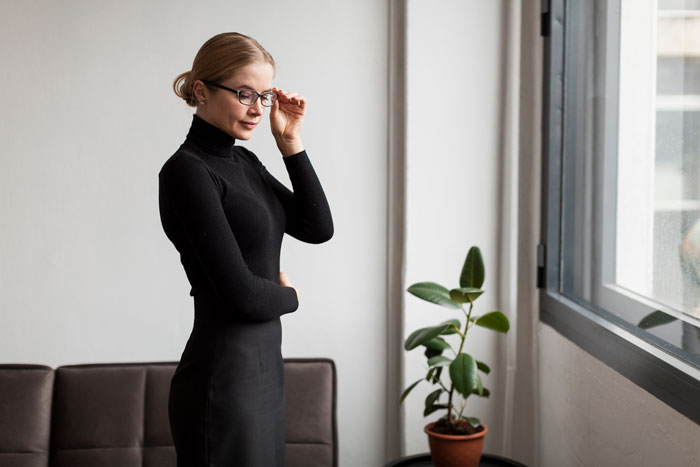 Woman wearing glasses and a black turtleneck standing near a window, reflecting on controlling behavior and clapping back.