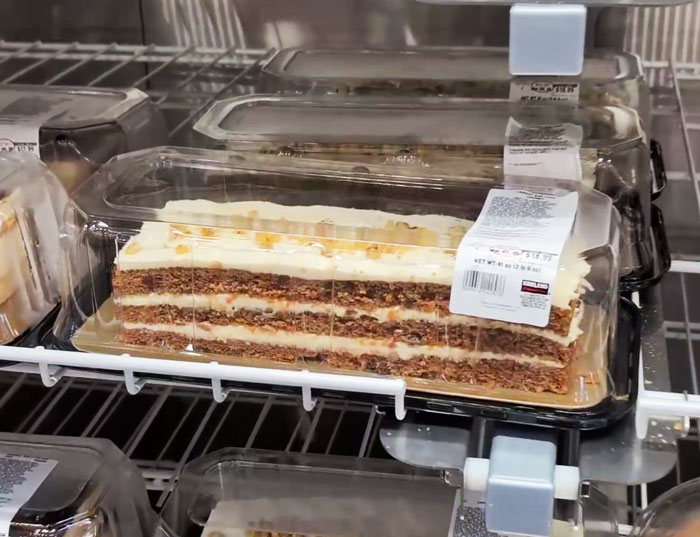 Costco carrot cake slice in plastic container on store shelf, featuring layers and white frosting.