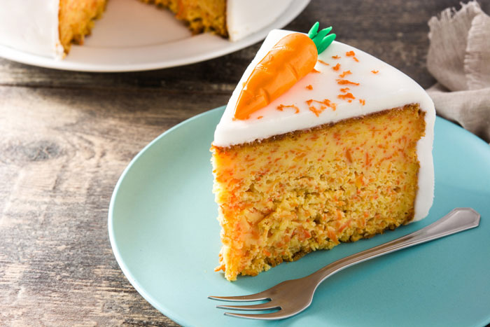 Slice of carrot cake on a blue plate with icing and garnished with a fondant carrot, sparking debate among Costco shoppers.