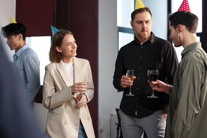Coworkers in party hats discuss a gluten-free diet while holding drinks, illustrating personal sharing at work events.