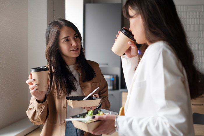 Two coworkers discussing gluten-free diets while holding coffee and salads in an office break room.