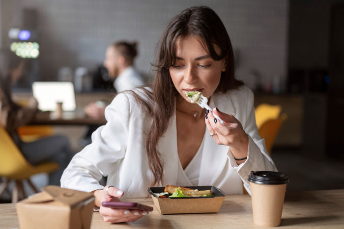 Woman in a white blazer eating a gluten-free meal at a desk, holding a phone, with coffee nearby.