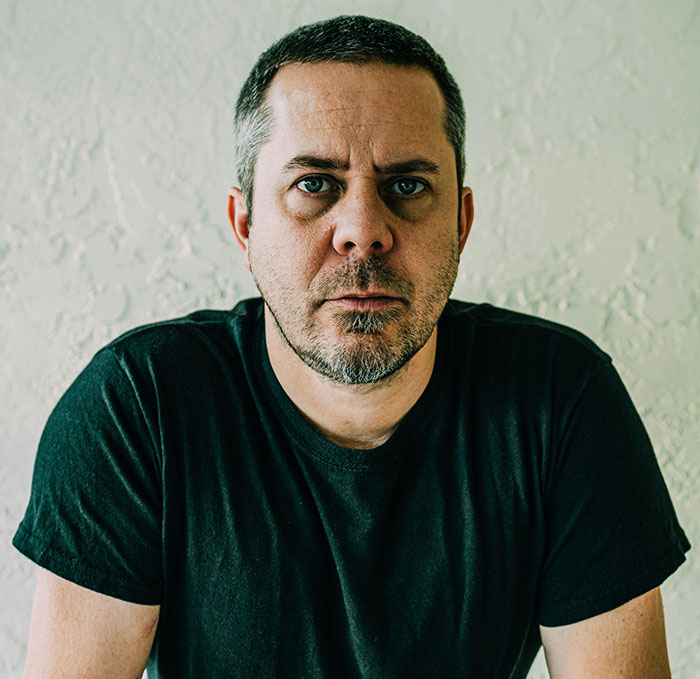 Man in a black shirt with a serious expression, in front of a textured wall, illustrating a client's delusional state.