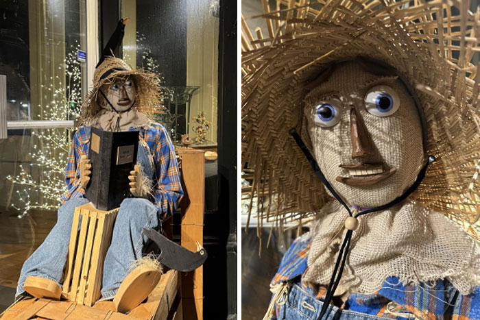 Creepy scarecrow figure wearing a straw hat and plaid shirt, sitting and holding a book in a dimly lit setting.