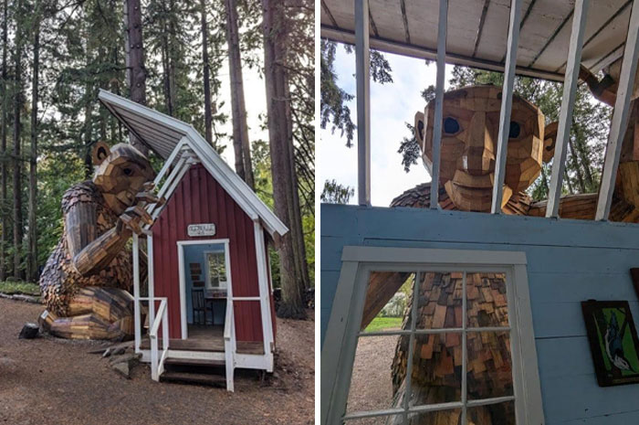 Large creepy wooden troll sculpture peering into a small red cabin in a forest setting, evoking eerie and unsettling vibes.