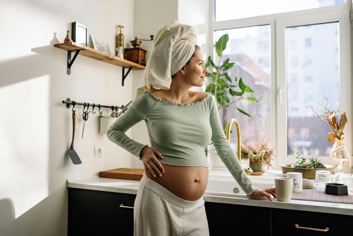 Pregnant woman in a kitchen, hand on hip, wearing a towel on her head, near a window and plants.