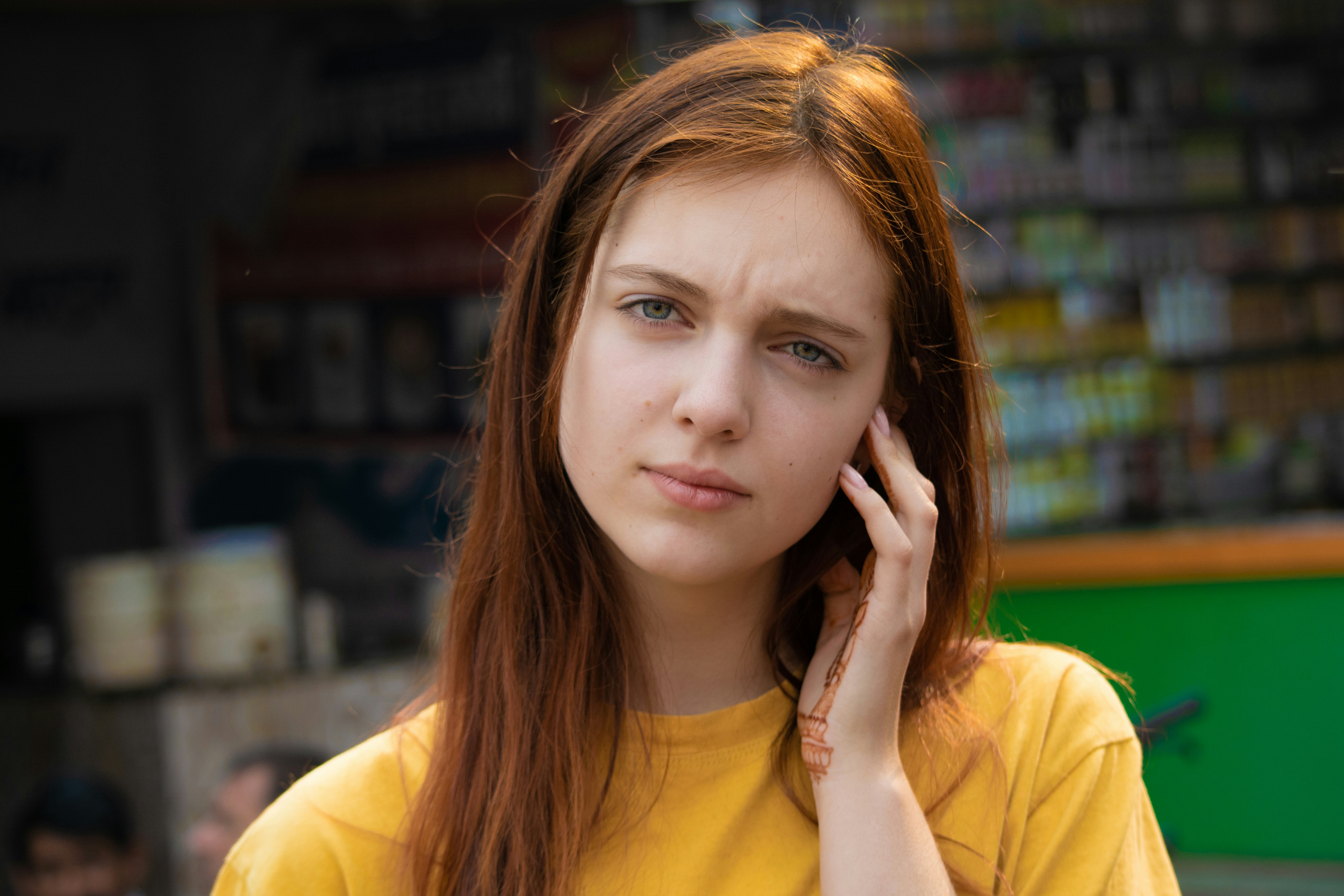 Young woman in a yellow shirt looking thoughtful, touching her face outdoors.