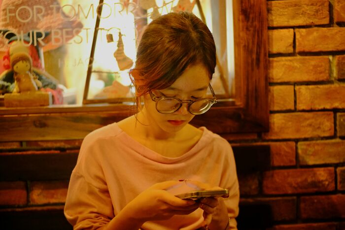 Young woman in glasses sharing red pill stories on her smartphone in a cozy cafe with warm lighting.