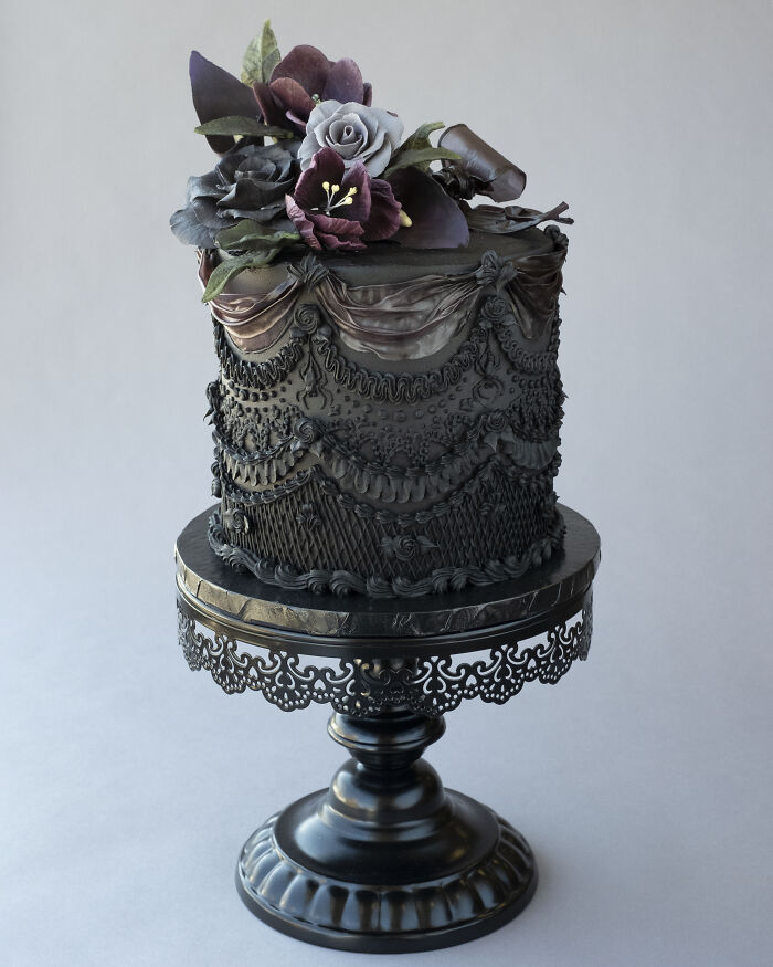 Vegan cake decorated with intricate lace designs and dark flowers on an ornate cake stand.