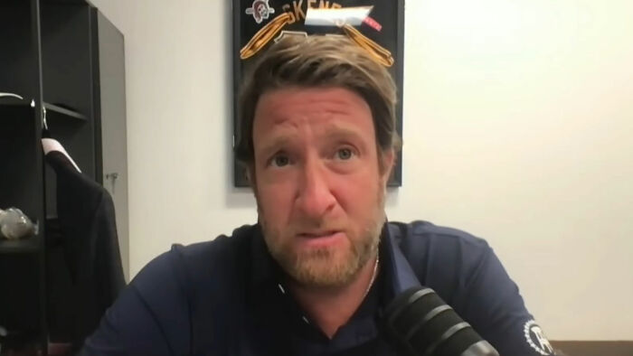 A person in a blue shirt speaking into a microphone, framed sports memorabilia in the background.