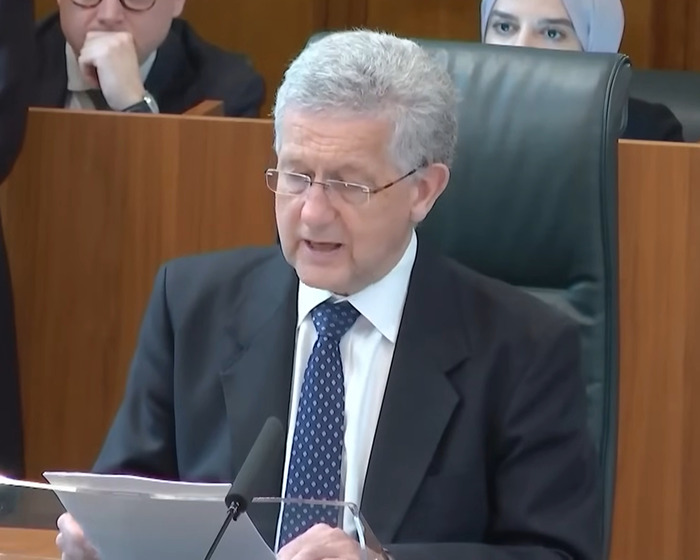 Man in a suit and glasses speaking at a podium, related to David Tennant trans allyship discussion.