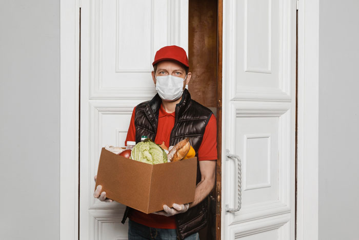 Delivery driver in a red cap and mask holding a grocery box at an open front door. Delivery driver in a red cap and mask holding a grocery box at an open front door.